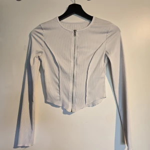 Vit ribbad långärmad tröja med dragkedja - Säljer en vit ribbad långärmad tröja med dragkedja framtill. Tröjan är croppad med lätt vågig nederkant och har en tight passform. Perfekt för dig som gillar stilrena och trendiga plagg med snygga detaljer. Använd 2 gånger 