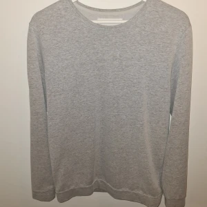 Ljusgrå basic tröja från Samsøe Samsøe - En stilren ljusgrå långärmad tröja från Samsøe Samsøe i mjuk bomullsmix. Enkel design med rund halsringning och normal passform, perfekt för en clean och avslappnad look. Diskret logga i sidan. Passar till allt och är superbekväm.