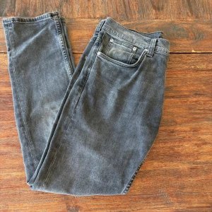 Grå jeans från Nudie Jeans, W31 L30 - Säljer ett par grå jeans från Nudie Jeans med klassisk femficksdesign och diskret broderad bakficka. Jeansen har normal passform och raka ben, tillverkade i mjuk bomullsdenim. Perfekta för dig som gillar stilrena och avslappnade vibes.