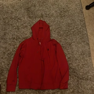 Röd hoodie från Polo Ralph Lauren - Röd hoodie från Polo Ralph Lauren med klassisk huva och snörning. Tröjan har lång ärm, normal passform och liten broderad logga på bröstet. Perfekt för chill dagar eller när du vill ha en enkel men snygg look.