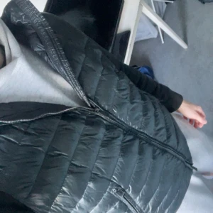 Svart dunväst med huva - Snygg svart dunväst med quiltad design och dragkedja framtill. Västen har en praktisk huva och en stilren, mörk look som passar perfekt över en hoodie. Materialet är glansigt och ger en modern vibe. Perfekt för dig som gillar streetwear och vill hålla värmen.