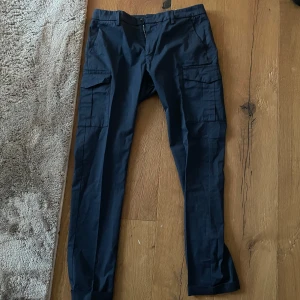 Dondup, stl 30 - Snygga mörkblå cargobyxor från Dondup med raka ben och flera praktiska fickor på sidorna. Byxorna har klassisk cargostil och är tillverkade i ett slitstarkt bomullsmaterial. Perfekta för en avslappnad och trendig look.