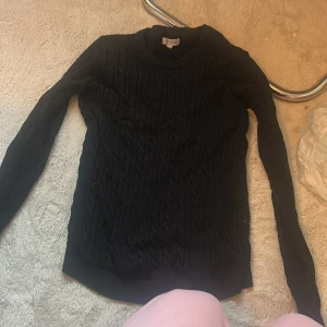 Svart kabelstickad tröja - Svart kabelstickad tröja med lång ärm och rund hals. Tröjan har ett klassiskt flätmönster och är gjord i ett mjukt stickat material som passar perfekt när det är kyligt ute. Enkel och stilren modell som är lätt att matcha med olika outfits.