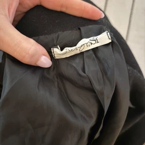 Svart kort kappa från Design by Si - Stilren svart kort kappa med dubbelknäppning och bred krage från Design by Si. Jackan har en rak och lite loose passform, långa ärmar. Perfekt för dig som gillar klassisk och enkel stil.🤩