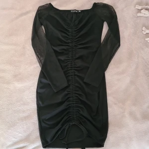 Svart meshklänning PrettyLittleThing - Svart kort klänning från PrettyLittleThing med långa meshärmar och snygg rynkdetalj framtill. Klänningen har snörning nertill och en tight passform som framhäver figuren. Perfekt för dig som gillar trendiga och stilrena plagg.