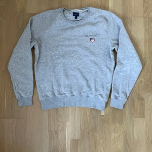 Grå sweatshirt från GANT i bomull - Snygg grå sweatshirt från GANT i storlek M. Tröjan har rund hals, ribbade muddar och ett broderat GANT-logo med sköld på bröstet. Tillverkad i mjuk bomull, perfekt för chill dagar. Enkel och stilren design som funkar till jeans eller joggers.