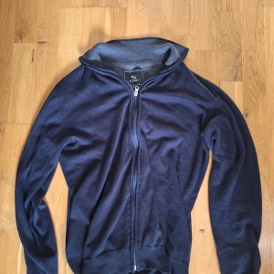 Mörkblå kofta från Earl William - Snygg mörkblå kofta från Earl William i storlek M. Tröjan har hel dragkedja framtill, ribbade muddar och hög krage. Perfekt för lager-på-lager och chill stil. Materialet är mjukt och stickat, vilket gör den skön att bära under kyliga dagar.