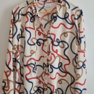 Mönstrad skjorta från Bondelid, strl 38 - Snygg skjorta från Bondelid i vitt med repmönster i blått, rött och brunt. Skjortan har långa ärmar, knappar framtill och dekorativ snörning vid halsen. Materialet känns lätt och flowy, perfekt för dig som gillar unika prints.