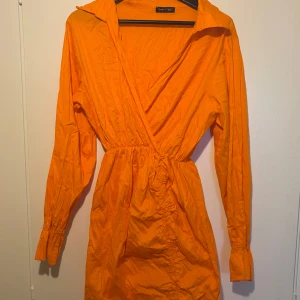 Orange klänning - Snygg orange kortklänning med omlott-design och v-ringning.  Klänningen har långa ärmar med resår vid handlederna och markerad midja. 