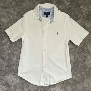 Vit kortärmad skjorta Polo Ralph Lauren - Säljer min sons vita kortärmad skjorta från Polo Ralph Lauren med klassisk krage och knappar framtill. Broderad logga på bröstet och insida krage med blåvita ränder. Skjortan är i bomull och har en clean, tidlös look.