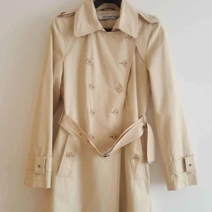 Beige trenchcoat från Easy Comfort - Stilren beige trenchcoat från Easy Comfort med klassisk dubbelknäppning, bälte i midjan och snygga detaljer vid ärmslut. Jackan har långa ärmar, bred krage och är tillverkad i ett bomullsliknande material. Perfekt för dig som vill ha en tidlös look.