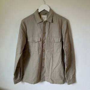 Beige overshirt från A Day's March - Stilren beige overshirt från A Day's March i 100% bomull. Skjortan har två bröstfickor med lock, knappar framtill och klassisk krage. Perfekt lager-på-lager-plagg med lång ärm och enkel, clean look. 2L2