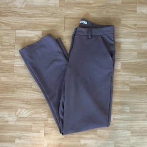 Bruna lågmidjade kostymbyxor från Lager 157 - Snygga lågmidjade bruna kostymbyxor från Lager 157 i stretchigt material med 95% polyester och 5% elastan. Byxorna har raka ben, bälteshällor och stängs med knapp och dragkedja. Perfekta för en clean och stilren look.