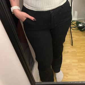 Svarta lågmidjade kostymbyxor från Lager 157 - Svarta lågmidjade kostymbyxor från Lager 157 med klassisk rak passform och diskreta fickor. Byxorna har hällor för bälte och stängs med knapp och dragkedja. Materialet är en mix av polyester, viskos och elastan vilket ger en mjuk och stretchig känsla.