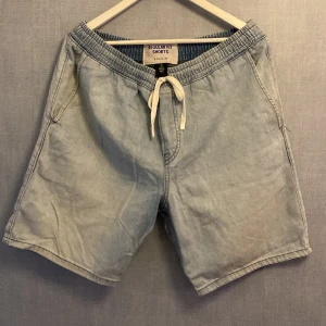 Ljusblå denimshorts från &Denim - Säljer ett par ljusblå denimshorts från &Denim i regular fit. Shortsen har elastisk midja med vit snörning, två sidofickor och en bakficka. Perfekta för sommardagar och har en avslappnad, loose passform.
