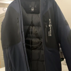 Sail Racing Pole Down Jacket navy - Säljer en mörkblå och svart dunjacka från Sail Racing, modell Pole Down Jacket. Jackan har Gore-Tex-yttermaterial, flera dragkedjeförsedda fickor och hög krage med huva. Perfekt för kalla dagar och har en clean, sportig look.