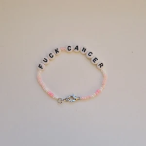 Pärlarmband med text Fuck Cancer - Armband med vita runda pärlor som stavar ut 'FUCK CANCER' i svarta bokstäver. Resten av armbandet består av små pärlor i ljusrosa och vita nyanser. Stängs med en silvrig karbinhake. Perfekt statement-piece för att visa stöd och styrka både att ge bort eller behålla själv. Storleken är ungefär 16-17 cm men är också gjorde med töjbar tråd🩷