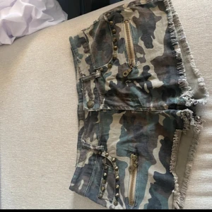Camo shorts med nitar och dragkedjor - Säljer ett par riktigt snygga camo-mönstrade shorts med råa kanter, dragkedjor och coola nitar framtill. Shortsen har fickor både fram och bak samt bälteshällor. Perfekta för dig som vill sticka ut med en edgy streetstil.