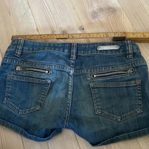 Blå jeansshorts med dragkedjor Five 5PM - Snygga blå jeansshorts från Five 5PM med coola dragkedjor och nitar på fickorna. Klassisk femficksmodell med uppvikta ben och midwaist passform. Perfekt för dig som gillar en avslappnad och trendig stil på sommaren.