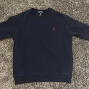 Mörkblå sweatshirt från Polo Ralph Lauren - Klassisk mörkblå sweatshirt från Polo Ralph Lauren med rund halsringning och röd broderad logga på bröstet. Tröjan har långa ärmar och en stilren, enkel design som passar perfekt till jeans eller chinos.