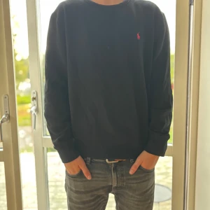 Svart sweatshirt från Polo Ralph Lauren - Svart sweatshirt från Polo Ralph Lauren med klassisk röd broderad logga på bröstet. Tröjan har rund hals, långa ärmar och är tillverkad i mjuk bomull. Perfekt för dig som gillar stilrena och enkla plagg med en touch av premiumkänsla.