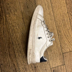 Vita sneakers från Polo Ralph Lauren - Snygga vita sneakers från Polo Ralph Lauren med klassisk logga på sidan och Polo-tryck på snörningen. Skorna har låg profil, rund tå och platt sula. Ovandelen är i skinn med mörkblå detalj på hälen. Perfekta för en clean och sportig stil.