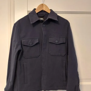 Mörkblå overshirt från Zara - Stilren mörkblå overshirt från Zara med klassisk krage, två stora bröstfickor med lock och knappar samt raka ärmar med knappdetalj vid mudden. 