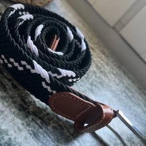Svart och vit flätad textilbälte - Snyggt flätat bälte i svart och vitt med bruna skinn-detaljer vid spännet. Bältet har ett randigt mönster och är gjort av slitstark textil med inslag av skinn. Perfekt för att ge outfiten en sportig och stilren touch.