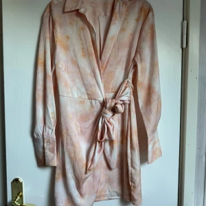 Omlott kortklänning i ljusa toner - Säljer en snygg omlottklänning med knyt i midjan och krage. Klänningen har ett unikt batikmönster i ljusa champagnefärger och långa ärmar med manschett. Perfekt för dig som gillar trendiga detaljer och mjuka färger.