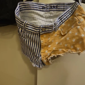 Randiga och stjärnmönstrade shorts - Unika shorts från KSUBI med ena sidan blåvit randig och andra sidan gul med vita stjärnor. Råa kanter nertill ger en cool, avslappnad vibe. Klassisk femficksmodell med knapp och dragkedja. Perfekta för dig som vill sticka ut i sommar!