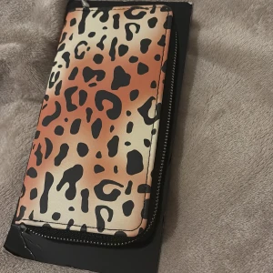 Leopardmönstrad plånbok från Woolworth - Cool plånbok med leopardmönster i beige, brunt och svart. Plånboken har dragkedja runtom och flera fack inuti, inklusive myntfack med egen dragkedja. Tillverkad i syntetmaterial och perfekt för dig som vill sticka ut med din accessoar.