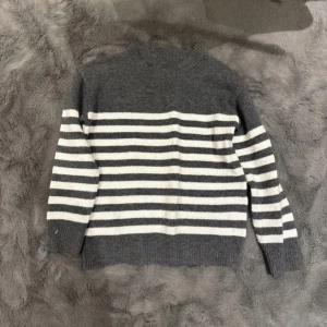 Randig grå och vit stickad tröja - Mysig stickad tröja i grått och vitt med klassiskt horisontellt randigt mönster. Tröjan har rund halsringning och långa ärmar, perfekt för kyliga dagar. Passar dig som gillar enkel men stilren look.