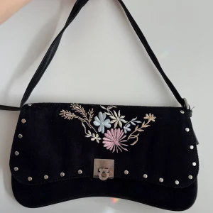 Svart axelväska med broderade blommor - Svart axelväska i mocka med broderade blommor i pastellfärger på locket. Väldigt fin och vintage. Skriv om du har frågor💗