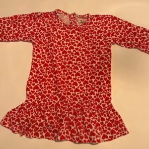 Röd hjärtklänning med volangkant - Supersöt kort klänning från Åhléns med rött hjärtmönster på vit botten. Klänningen har långa ärmar och en volangkant nertill. Tillverkad i mjuk bomull och modal, perfekt för små fashionistas som gillar färg och mönster. Storlek 74/80