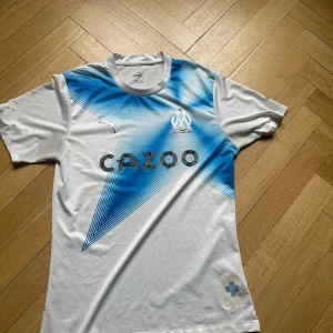 Marseille fotbollströja Puma vit/blå - Säljer en Olympique de Marseille fotbollströja från Puma. Tröjan är vit med ett stort blått grafiskt mönster framtill, klubbmärke och sponsortryck. Tillverkad i lätt och ventilerande polyester, perfekt för match eller träning.