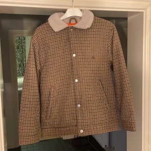 Rutig jacka från Filippa K med teddykrage - Snygg rutig jacka från vailentmed beige och bruna toner, mjuk teddykrage och orange quiltat foder. Jackan har tryckknappar framtill, två fickor och lång ärm. Perfekt för höst och vinter när du vill ha både stil och värme.