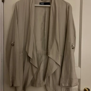 Beige tunn jacka  från Zara - Snygg beige tunn jacka från Zara med avslappnad passform och vattenfallsframsida. Jackan har långa ärmar med dekorativa metallringar där man kan vika upp ärmarna och är tillverkad i ett mjukt, lätt tyg som ger en stilren look.