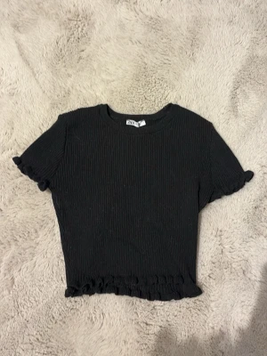 Svart croppad topp från ZARA - Säljer en svart croppad topp från ZARA med ribbad struktur och volangdetaljer vid ärmslut och nederkant. Toppen har korta ärmar och en rund halsringning, perfekt för en trendig och enkel look.