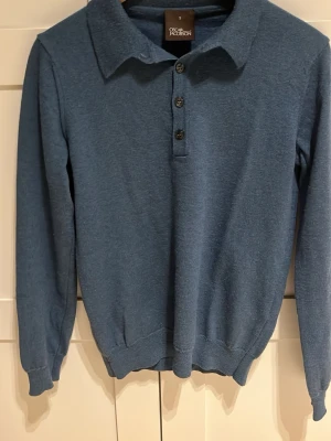 Merino ull tröja från Oscar Jacobson - Snygg blå långärmad tröja från Oscar Jacobson med krage och tre knappar framtill. Tröjan är gjord av merinoull. Tröjan har ribbade muddar vid ärmslut och nederkant, vilket ger en clean och stilren look. Perfekt för dig som gillar klassisk men avslappnad stil. Tröjan är i storlek S men sitter som Xs.