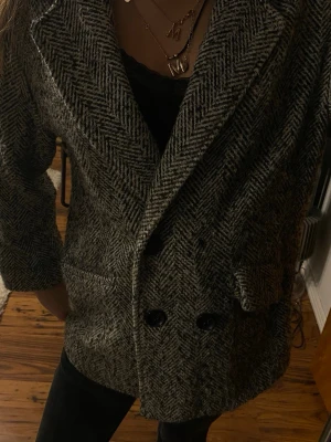 Grå kavaj med fiskbensmönster - Snygg grå kavaj med klassiskt fiskbensmönster och breda slag. Dubbelknäppt framtill med svarta knappar och två fickor. Perfekt oversized look som passar till allt. Materialet känns tjockt och mjukt, troligen ullblandning.