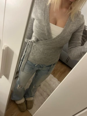 Grå omlottkofta från Gina Tricot - Mysig grå stickad omlottkofta från Gina Tricot med knytband i midjan och djup v-ringning. Koftan har långa ärmar och ribbade muddar. Perfekt att slänga över ett linne för en avslappnad men snygg look.