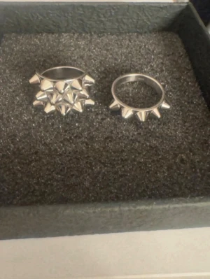 Två silverfärgade ringar med nitar Edblad  - Säljer två coola ringar i silverfärgad metall med spetsiga nitar runt hela ringen. Ena ringen har dubbla rader av nitar för extra edgy look, medan den andra har en enkel rad. Perfekta accessoarer för dig som gillar statement smycken storlek 17,5