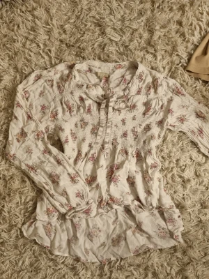 Blommig blus från Stories, M - Romantisk blommig blus från Stories i vitt med små rosa och beige blommor. Blusen har långärmad design, smockad överdel och knytband vid halsen. Tillverkad i ett lätt, mjukt tyg som ger en luftig känsla. Skulle säga att den passar mer som en 36/38 (S).