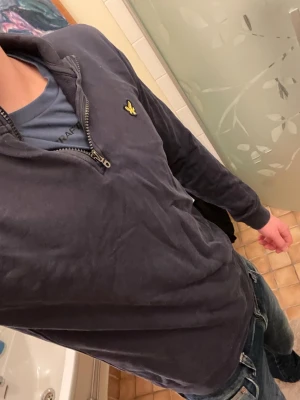 Mörkblå halvzip från Lyle & Scott - Snygg mörkblå tröja från Lyle & Scott med halv dragkedja och den klassiska gula loggan på bröstet. Tröjan har lång ärm och en avslappnad passform, perfekt att slänga på sig över en t-shirt. Tillverkad i mjukt bomullsmaterial.