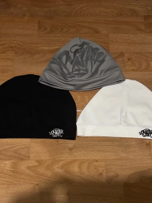 Tre mössor Syna World - Säljer tre mössor från Syna World i svart, vitt och grått. Alla har ett coolt streetstyle-inspirerat tryck framtill. Mössorna är mjuka och stretchiga, perfekta för dig som gillar enkel men snygg stil. Passar både till chill och mer urban look.