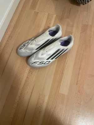 Adidas F50 vita fotbollsskor - Säljer ett par Adidas F50 fotbollsskor i vitt med svarta detaljer och klassiska tre ränder på sidan. Skorna har en strömlinjeformad design och är tillverkade i syntetmaterial för lätt känsla på planen. Perfekta för dig som vill ha snabbhet och stil på fotbollsplanen.