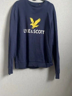 Marinblå sweatshirt från Lyle & Scott - Snygg marinblå sweatshirt från Lyle & Scott med gul broderad örn och vit logga på bröstet. Tröjan har rund halsringning, långa ärmar och ribbade muddar. Perfekt för en avslappnad och stilren look.Kan gå ner i pris 