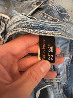 Ljusblå jeans från Nudie Jeans - Säljer ett par ljusblå jeans från Nudie Jeans med slitningar och hål på benen för en riktigt avslappnad och trendig look.