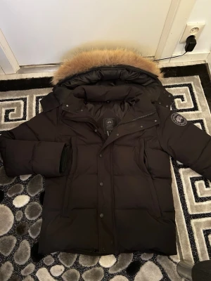 Svart dunjacka från Canada Goose - Snygg svart dunjacka från Canada Goose med stor huva och avtagbar pälskant. Jackan har klassisk patch på ärmen, dragkedja och knappar framtill samt flera praktiska fickor. Perfekt för kalla vinterdagar och riktigt varm.