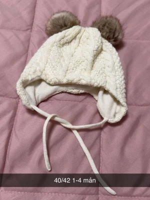 Vit stickad babymössa med tofsar - Supersöt vit stickad babymössa med två fluffiga bruna tofsar på toppen och knytband under hakan. Mössan har ett fint kabelstickat mönster och täcker öronen för extra värme. Perfekt för små barn under kalla dagar.